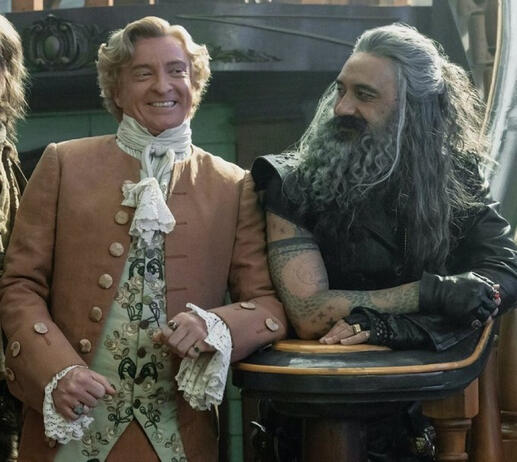 ed, in his black leather attire, stares at stead with a smile. Stede, smiling, is wearing an orange coat with large buttons and lacy sleeves.